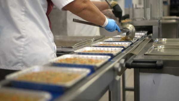 Gloved workers filling meal trays along a stainless-steel conveyor.